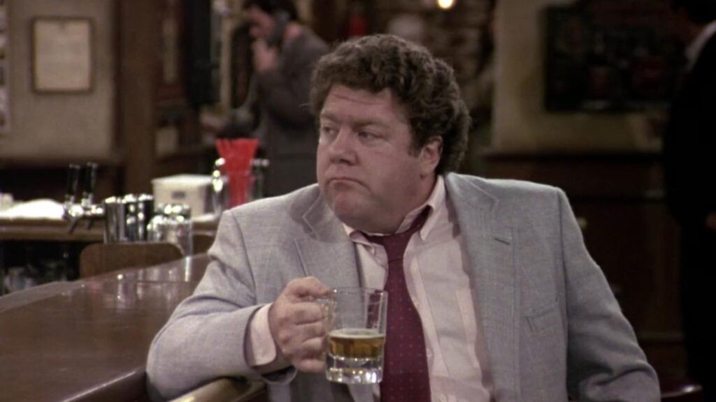 George Wendt Cheers as Norm Peterson sits at the Cheers bar with a beer, Sam Malone in the background.