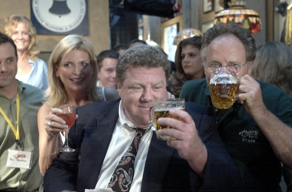 George Wendt Cheers as Norm Peterson with a beer, Cheers bar sign glowing, cast silhouettes cheering.